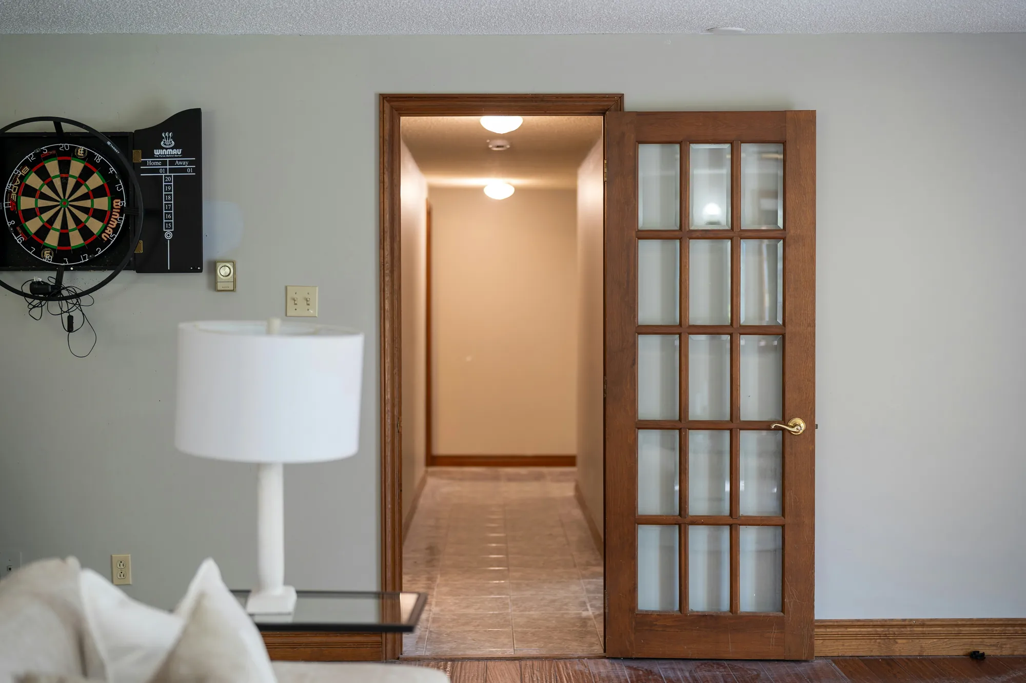 Finished basement game room with a dartboard, table lamp, and a glass-paned French door opening to a tiled hallway (representative photo for home inspections in New York City, NY)