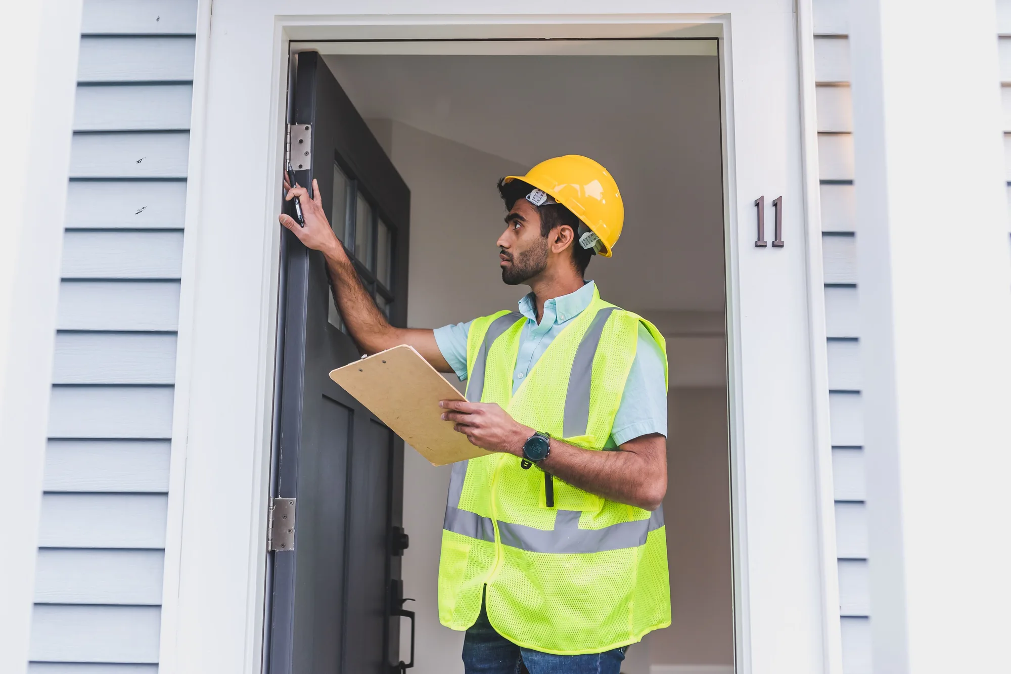 Home inspector in a hard hat and safety vest examining a front door hinge and frame while holding a clipboard, house numbered 11