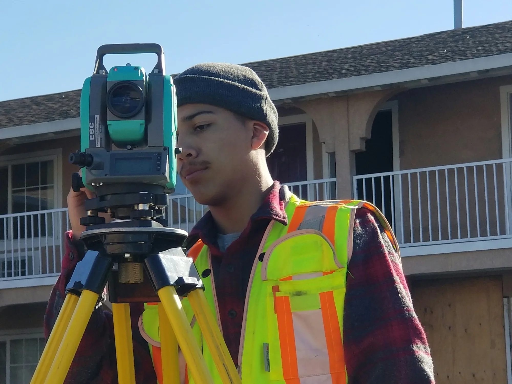 Surveyor in a safety vest and beanie using a total station survey instrument in front of a two-story multifamily apartment building during property due diligence