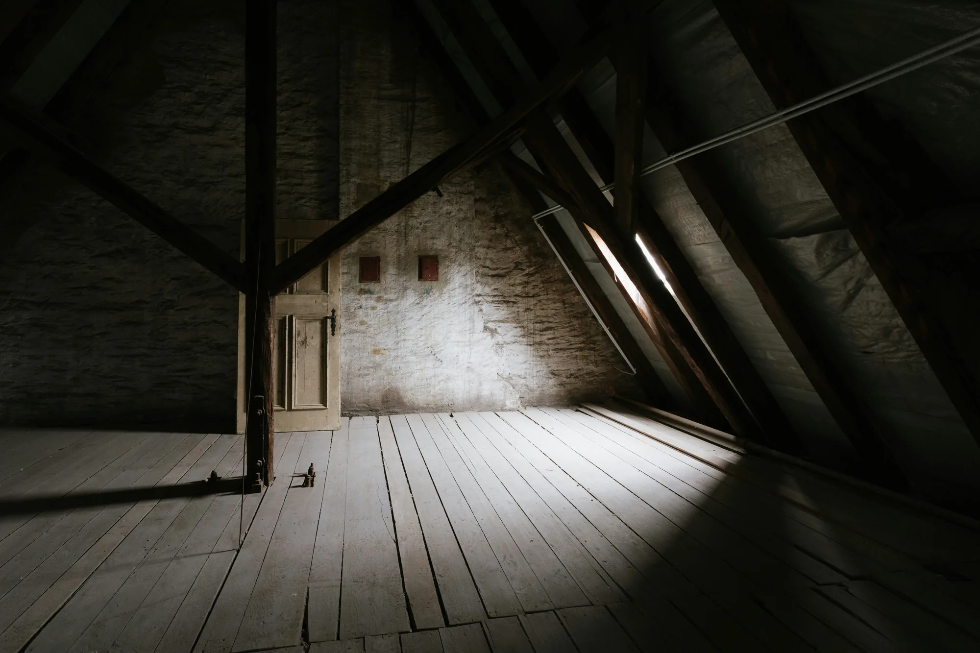 Dim unfinished attic space with exposed diagonal roof truss framing, a wood plank floor, and light coming through a small window, the kind of space where old wiring is often visible