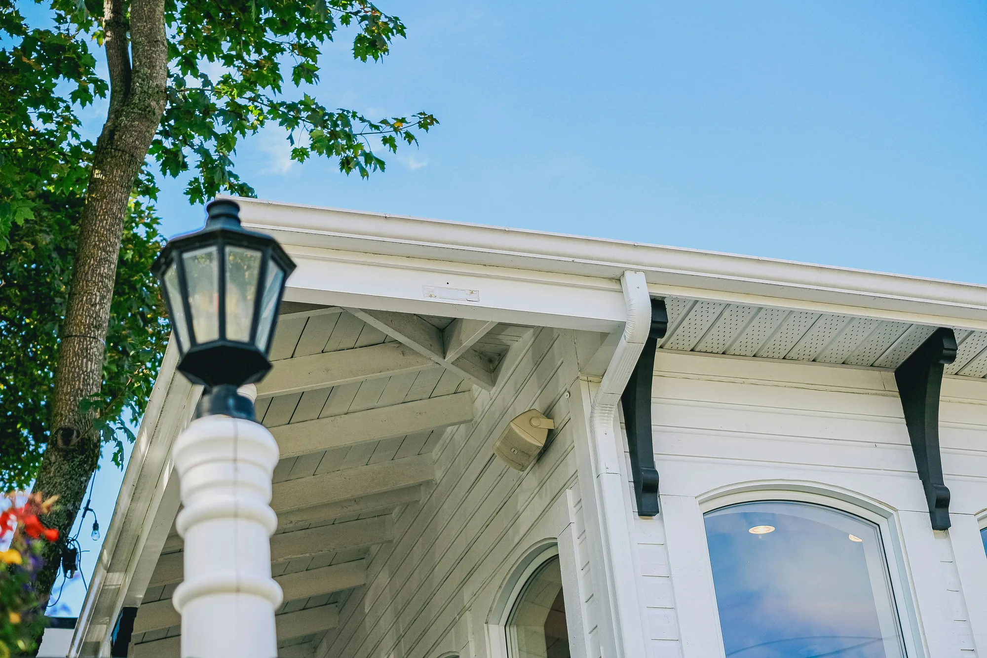 Close-up of a Victorian-style house's roofline, exposed rafter tails, gutter, and porch light against a clear blue sky (representative photo for home inspections in Broome County, NY)