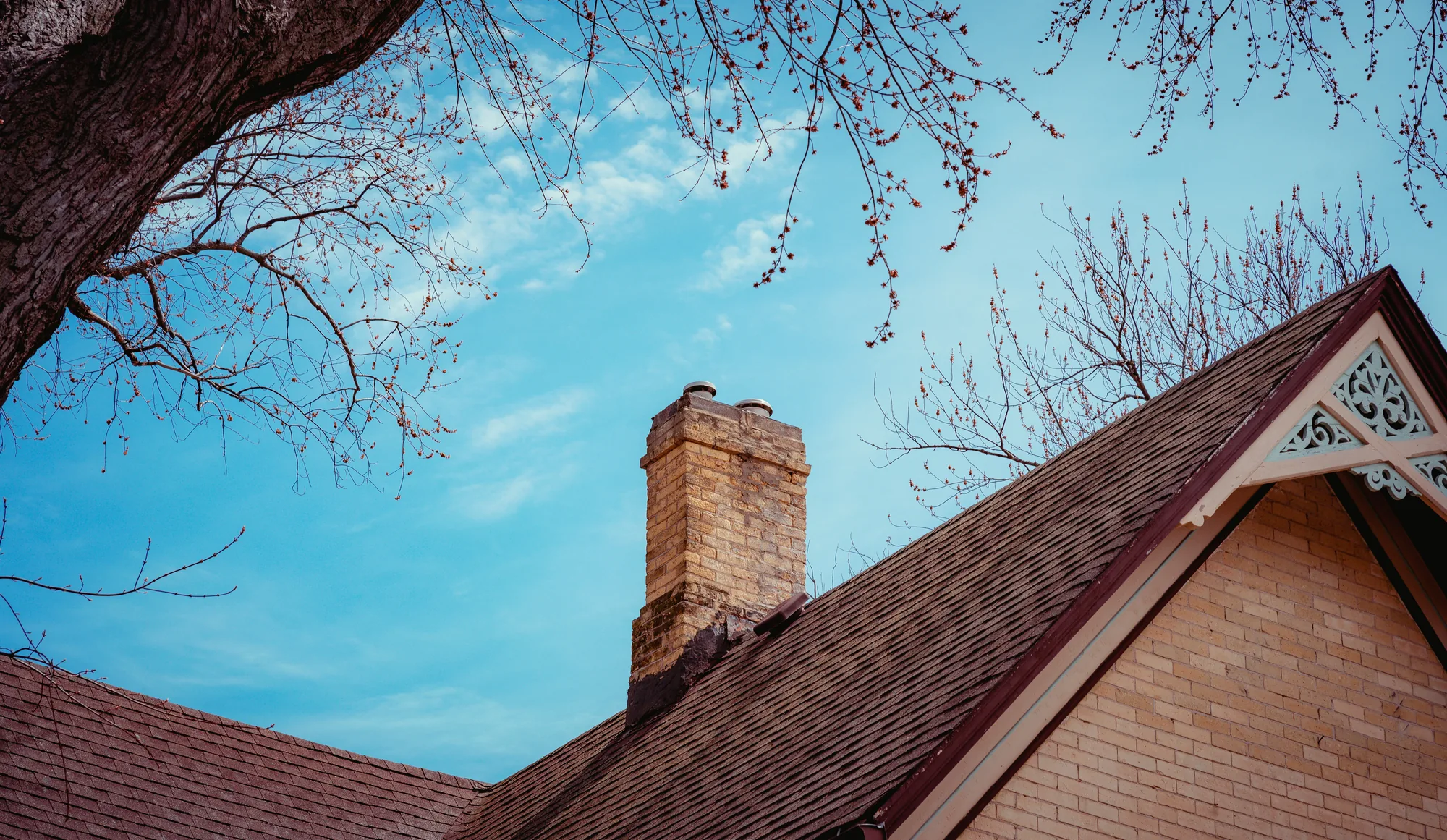 Asphalt shingle roof with a brick chimney and ornate white Victorian-style gable trim, framed by bare tree branches against a blue sky (representative photo for home inspections in Orleans County, NY)